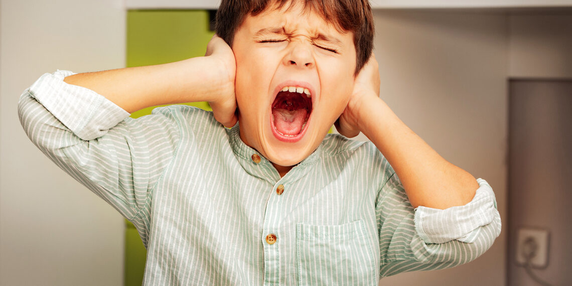 Little autistic boy with strong negative face expression closing ears and loudly screams