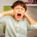 Little autistic boy with strong negative face expression closing ears and loudly screams