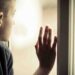 Close up Lonely Orphan Boy in an Orphanage Looking Outside While Holding the Glass Window.